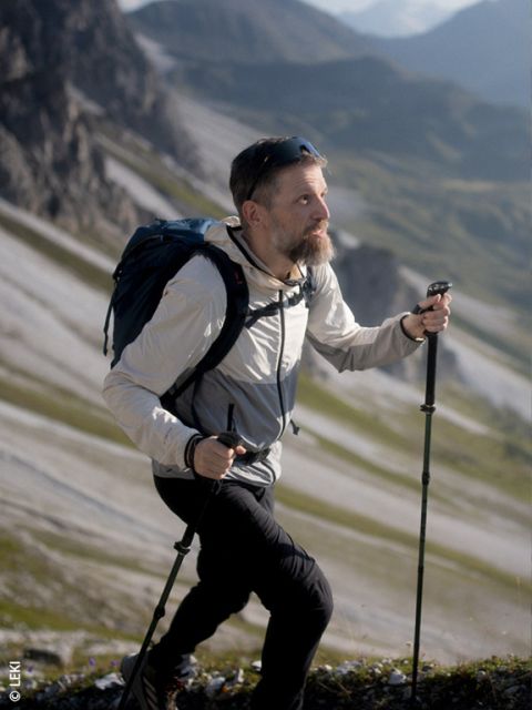 Un hombre con barba camina cuesta arriba por un sendero rocoso de montaña, usando bastones de senderismo y llevando una mochila; al fondo se ven montañas.