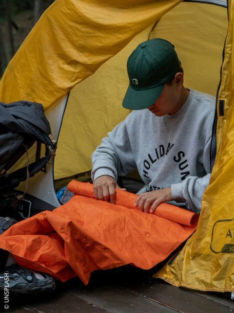 Una persona con un gorro verde está sentada en una tienda de campaña amarilla doblando una colchoneta naranja mientras acampa al aire libre.