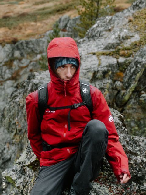 Una persona con una chaqueta roja con capucha y una mochila está sentada al aire libre sobre rocas, con árboles y terreno rocoso visibles al fondo.
