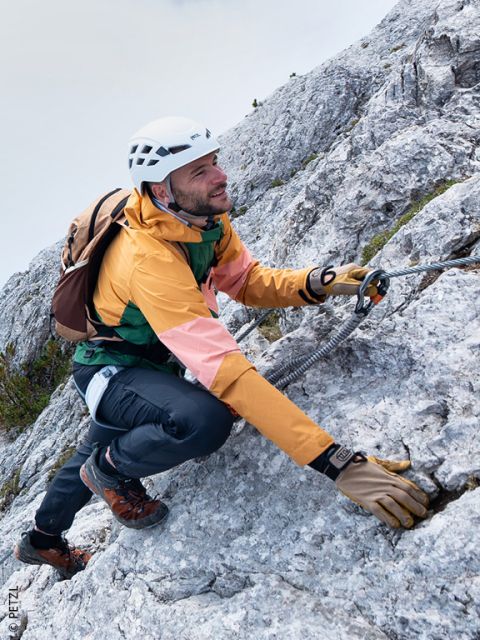 Un hombre con equipo de escalada sube por la ladera rocosa de una montaña, sujetándose a una cuerda de seguridad con un mosquetón.