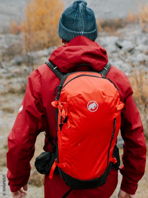 Una persona con una chaqueta roja y un gorro azul, haciendo senderismo en la naturaleza con una mochila naranja, vista desde atrás.