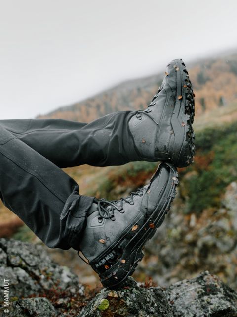 Una persona con botas de senderismo negras, con tierra y hojas adheridas, descansa sus pies al aire libre sobre rocas, con un paisaje montañoso borroso de fondo.