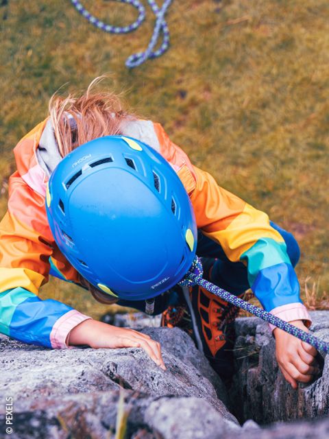 Una persona con casco azul y chaqueta colorida escala una pared de roca, asegurada por una cuerda. Vista desde arriba, debajo se ve un suelo cubierto de hierba.