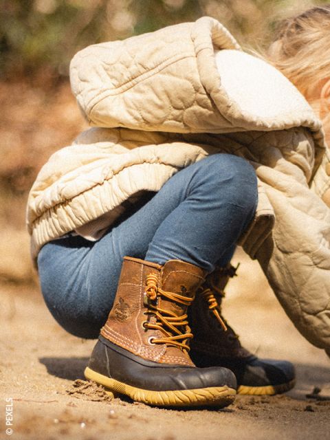 Un niño pequeño con un abrigo beige y botas marrones está en cuclillas al aire libre sobre un suelo arenoso, parcialmente de espaldas a la cámara.