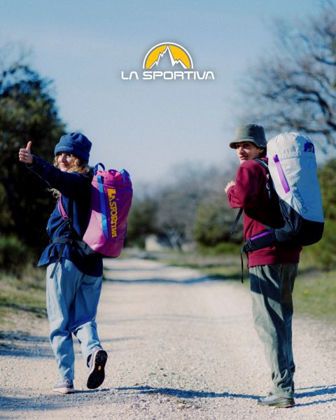 Cuatro personas con equipo de exterior y colchonetas de búlder a la espalda caminan por un sendero de montaña cubierto de hierba. Encima de ellos se ve el logotipo de La Sportiva en el cielo. Al fondo se ven escarpadas cumbres montañosas.