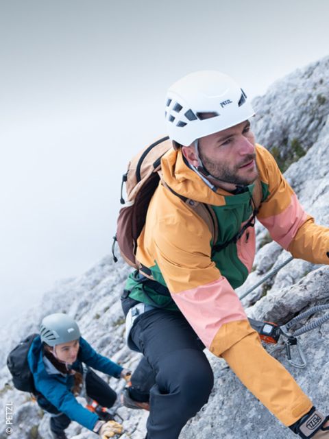 Un alpinista con casco, gafas de sol y equipo de escalada escala una ladera rocosa con picos nevados al fondo bajo un cielo despejado.