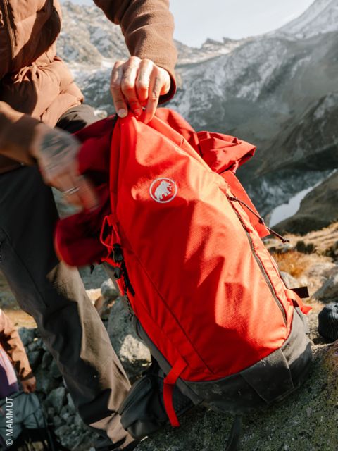 Una persona con ropa de invierno y guantes estampados pone una pala de nieve en una mochila rosa sobre un suelo cubierto de nieve, con árboles nevados al fondo.