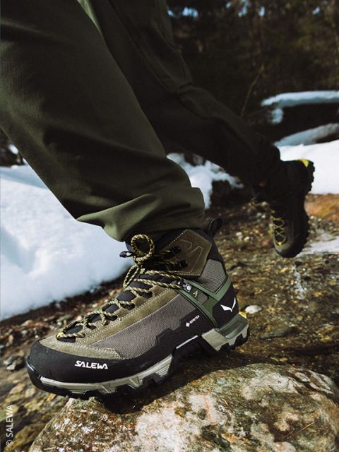 Una persona con ropa de exterior se ata los cordones de una bota de montaña negra mientras está de pie sobre una superficie rocosa, con una gran formación rocosa y un cielo azul de fondo.