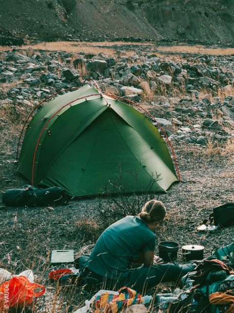 Una persona con chaqueta y gorro rojos prepara comida al aire libre, sentada en la hierba, cerca de equipo de camping, vista a través de la puerta abierta de una tienda de campaña con montañas y árboles al fondo.