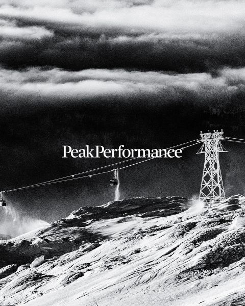 Una persona salta entre grandes rocas en una región montañosa bajo un cielo azul brillante. Lleva ropa deportiva, incluyendo una chaqueta y pantalones cortos. En la parte superior se puede ver la inscripción "Peak Performance".