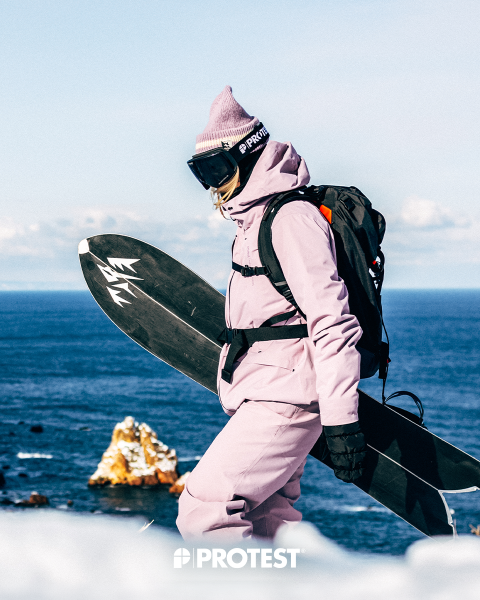 Una persona con un traje de nieve rosa y un gorro, sosteniendo una tabla de snowboard, está de pie junto al mar, con formaciones rocosas al fondo y la palabra "PROTESTA" debajo.