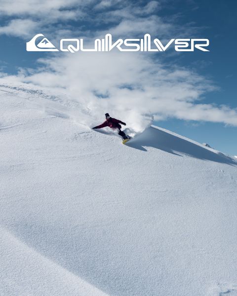 Un surfista cabalga una gran ola turquesa, personificando el equilibrio y la habilidad. Lleva pantalones cortos de baño oscuros, y la ola se encrespa sobre él. Encima de él se puede ver el logo de Quiksilver.