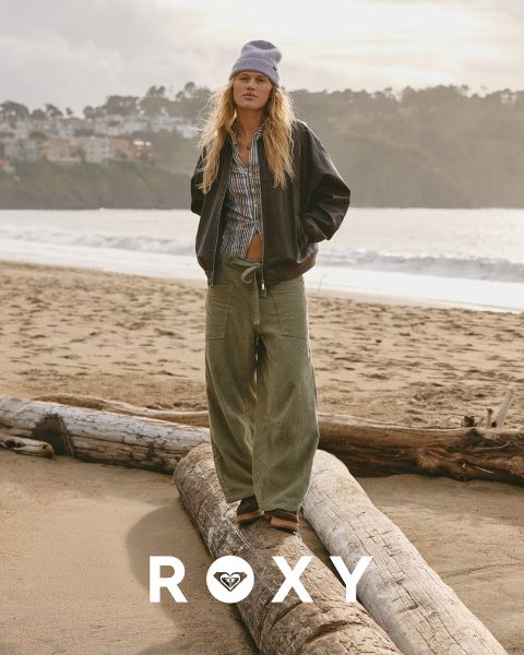 Tres mujeres nadan bajo el agua en trajes de baño en agua clara y azul. Una lleva una camisa de manga larga, las otras bikinis. La imagen está superpuesta con el logo blanco de "Roxy".
