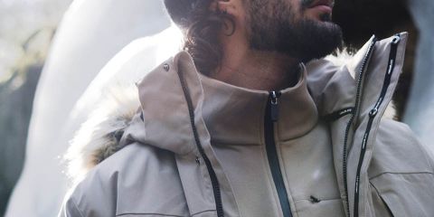 Hombre con chaqueta marrón, pantalones negros y gorro gris camina por un denso bosque. Lleva guantes negros y una mochila. El bosque tiene árboles altos y hojas caídas. Logotipo de "Rukka" en la parte superior.