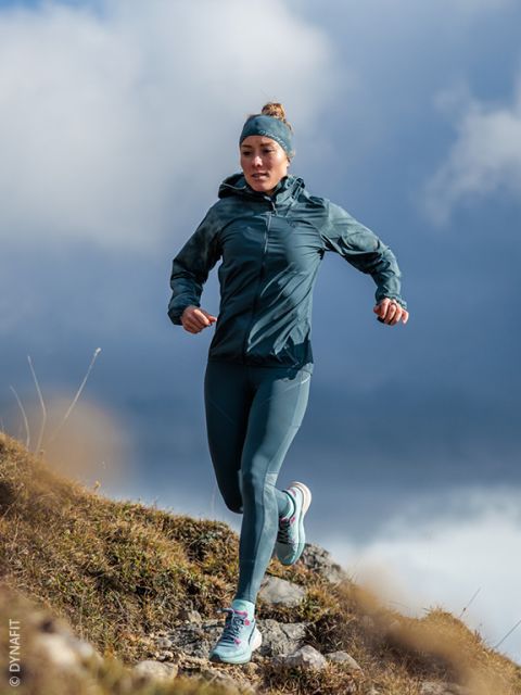 Una mujer con ropa deportiva turquesa corre cuesta abajo por un sendero rocoso, con un cielo nublado de fondo.