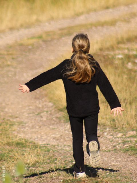 Una chica de pelo largo con un atuendo negro corre alegremente con los brazos extendidos por un camino de tierra bordeado de hierba.