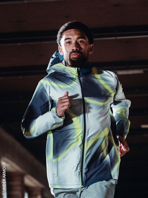 Una persona con una sudadera con capucha y zapatillas de correr trota por un sendero nevado en un día frío y despejado, con el horizonte de la ciudad al fondo.