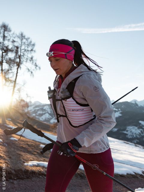Una mujer con una chaqueta y guantes rosa grisáceos corre en un paisaje montañoso nevado, llevando dos botellas de agua en los bolsillos delanteros de su chaleco para correr.