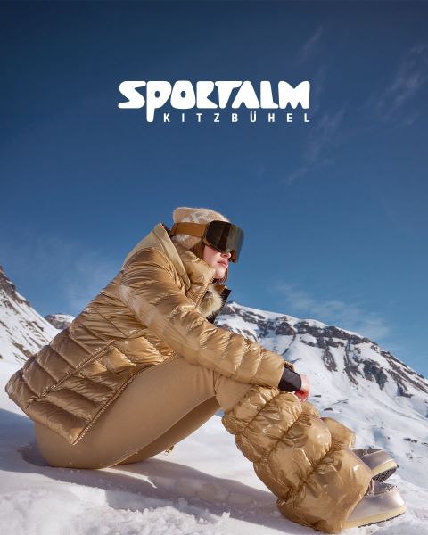 Una persona con una chaqueta acolchada metálica y pantalones negros yace en un terreno nevado con montañas al fondo bajo un cielo nublado. La escena es tranquila e invernal. En la esquina superior derecha se ve el texto "SPORTALM KITZBÜHEL".