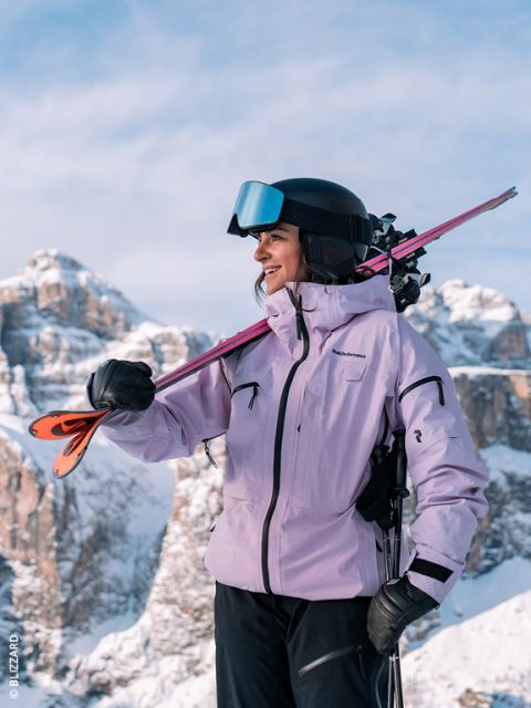 Una esquiadora con chaqueta lavanda y casco sonríe mientras lleva los esquís al hombro; al fondo se ven montañas nevadas.