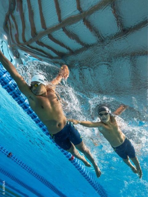 Dos nadadores con gafas y gorros de natación compiten bajo el agua en una piscina; uno extiende la mano hacia el otro cerca de la separación de carriles.