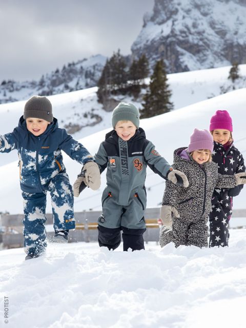 Cuatro niños con ropa de invierno juegan y sonríen en la nieve, con montañas y árboles cubiertos de nieve de fondo.