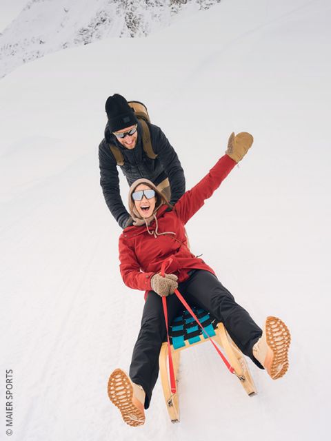 Dos personas con ropa de invierno tiran de trineos de madera cuesta arriba en una colina nevada, con árboles y una cima de montaña iluminada por el sol al fondo. Se pueden ver más personas en la distancia.
