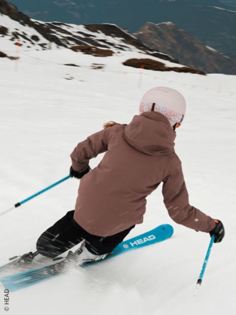 Un esquiador con una chaqueta beige, pantalones azules y un casco blanco desciende por una pista nevada, haciendo un giro brusco y levantando nieve. En el fondo se ven montañas.
