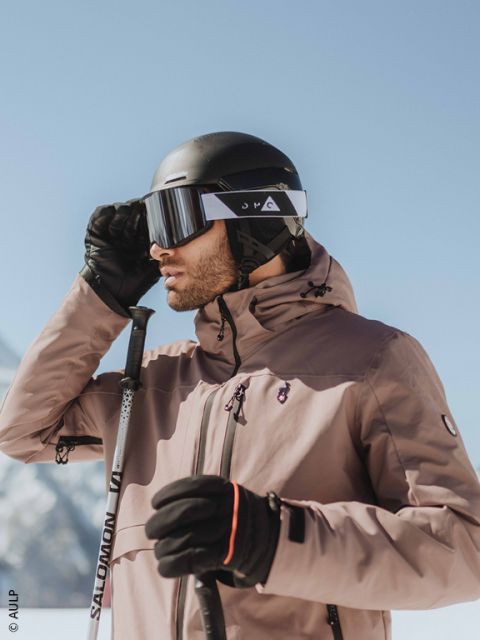 Primer plano de una persona con un casco de esquí gris, gafas de esquí reflectantes y una chaqueta oscura, con un paisaje nevado borroso al fondo.