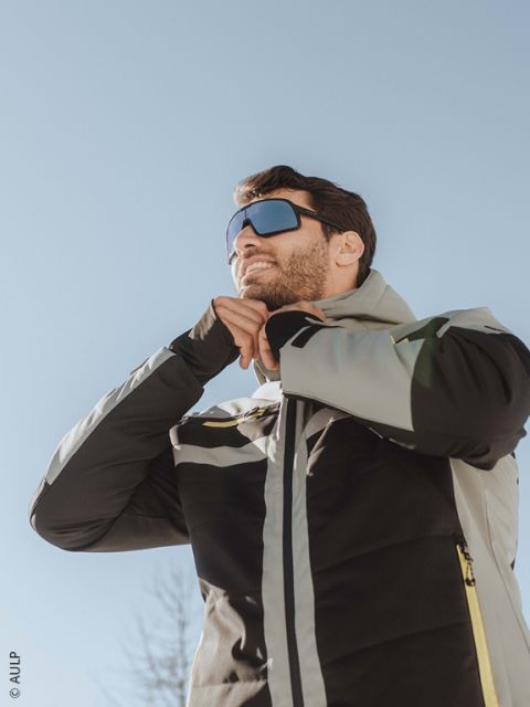 Un esquiador con casco blanco, gafas reflectantes, chaqueta marrón y negra y guantes se abrocha la chaqueta al aire libre con montañas nevadas al fondo.