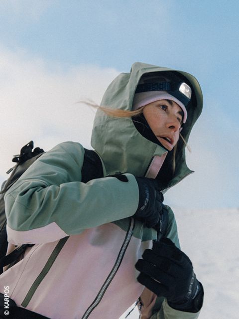 Una mujer, vestida con una chaqueta Dynafit acolchada, gafas de sol y guantes, se prepara para una caminata en un paisaje montañoso nevado, con picos cubiertos de nieve al fondo. Lleva una mochila y bastones de esquí.