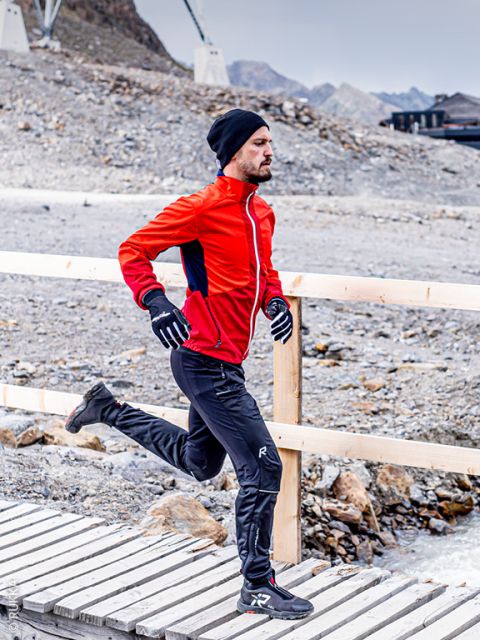 Un hombre con una chaqueta roja, pantalones negros, guantes y un gorro camina por un puente de madera en una zona rocosa y montañosa.