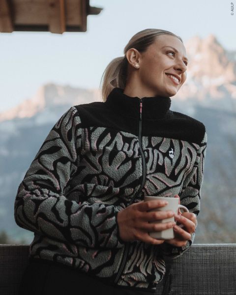Una mujer con una chaqueta polar estampada con cremallera sostiene una taza y mira las montañas al fondo.