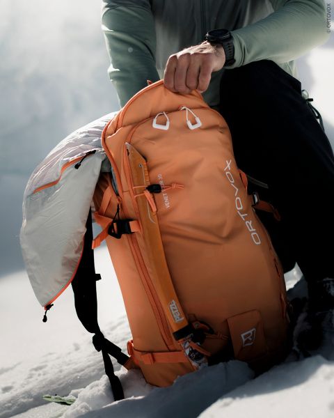 Un excursionista abre una mochila Ortovox naranja en un paisaje montañoso nevado.